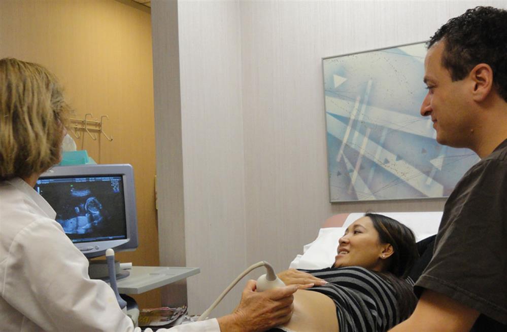 A health care provider holds an ultrasound probe against the abdomen of the pregnant woman lying on bed with her partner standing beside her while everyone looks at the ultrasound image on monitor.