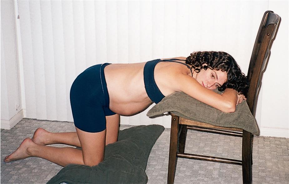 side close-up of a pregnant woman with head on the cushion above chair while knees are on the folded mattress above floor.