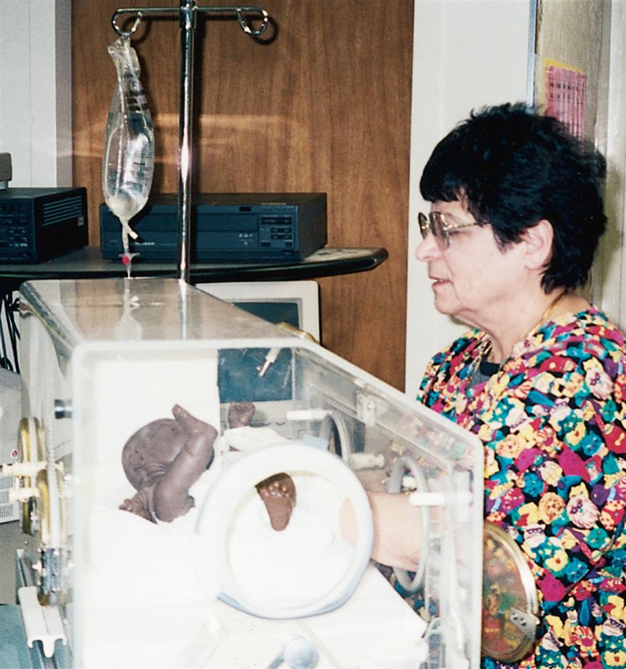Close-up of infant covered in diaper placed in incubator. A nurse holds the infant through window. An I V stand is on the one side of incubator.