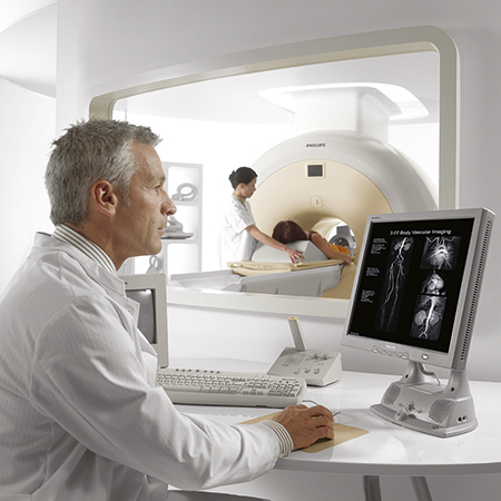 A magnetic resonance imaging technologist is viewing images on a monitor while a patient is lying on an x-ray table inside a scanner – a large machine with a hole in the center.