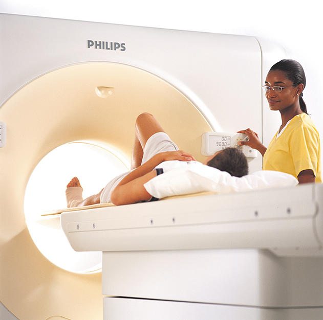A technologist is standing beside a patient lying on an x-ray table that slides into a CT scanner – a large machine with a hole in the center.